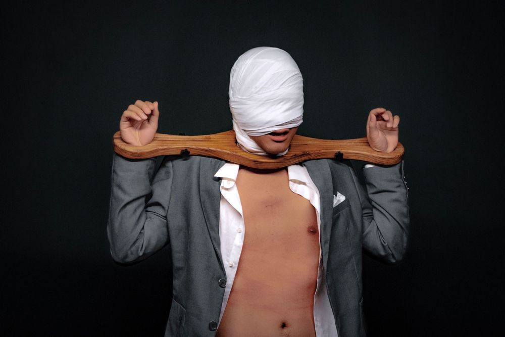 Man in gray suit with bandaged head holds wooden plank across chest against dark background.