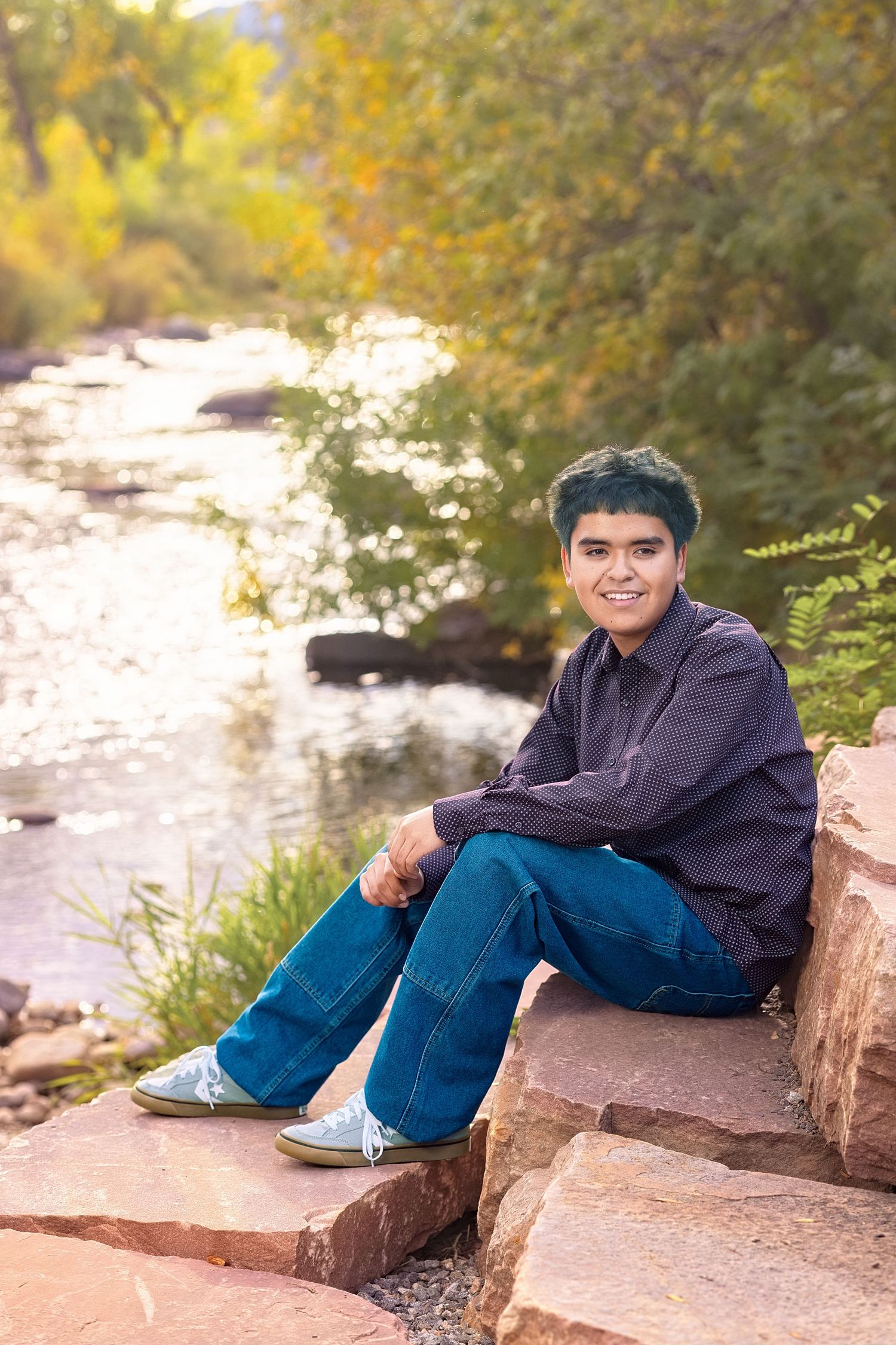 Teen with teal hair sits on red rocks by a shimmering creek, smiling in jeans and a dark shirt.