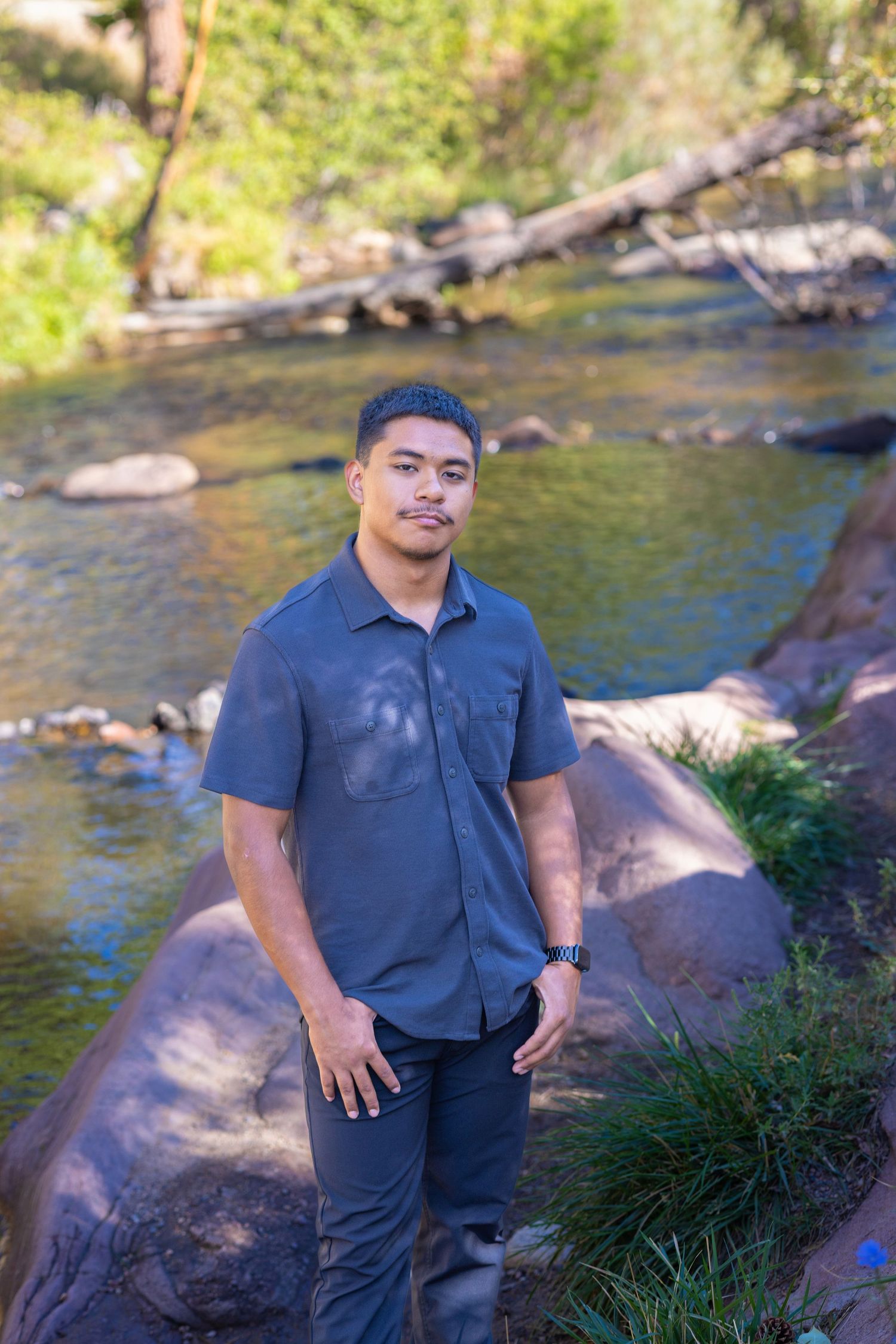 Young man in gray shirt poses on rocky riverbank with lush green trees and flowing stream in background.