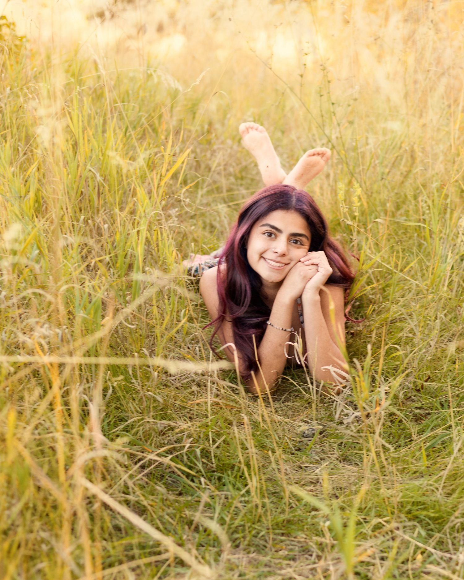 Young woman with dark hair lying in tall golden grass field, smiling peacefully in warm sunlight.