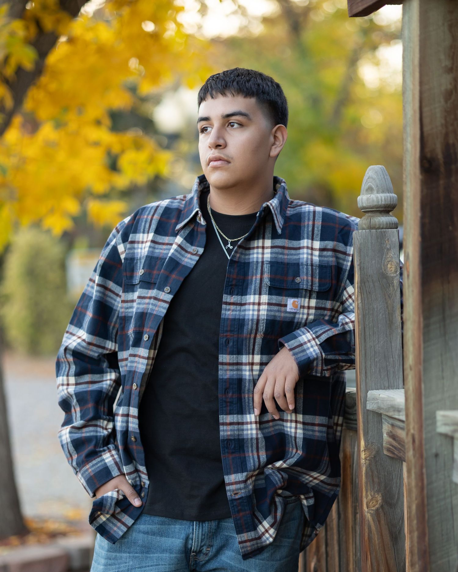 Young man in plaid flannel shirt leaning against wooden post with autumn yellow foliage background.