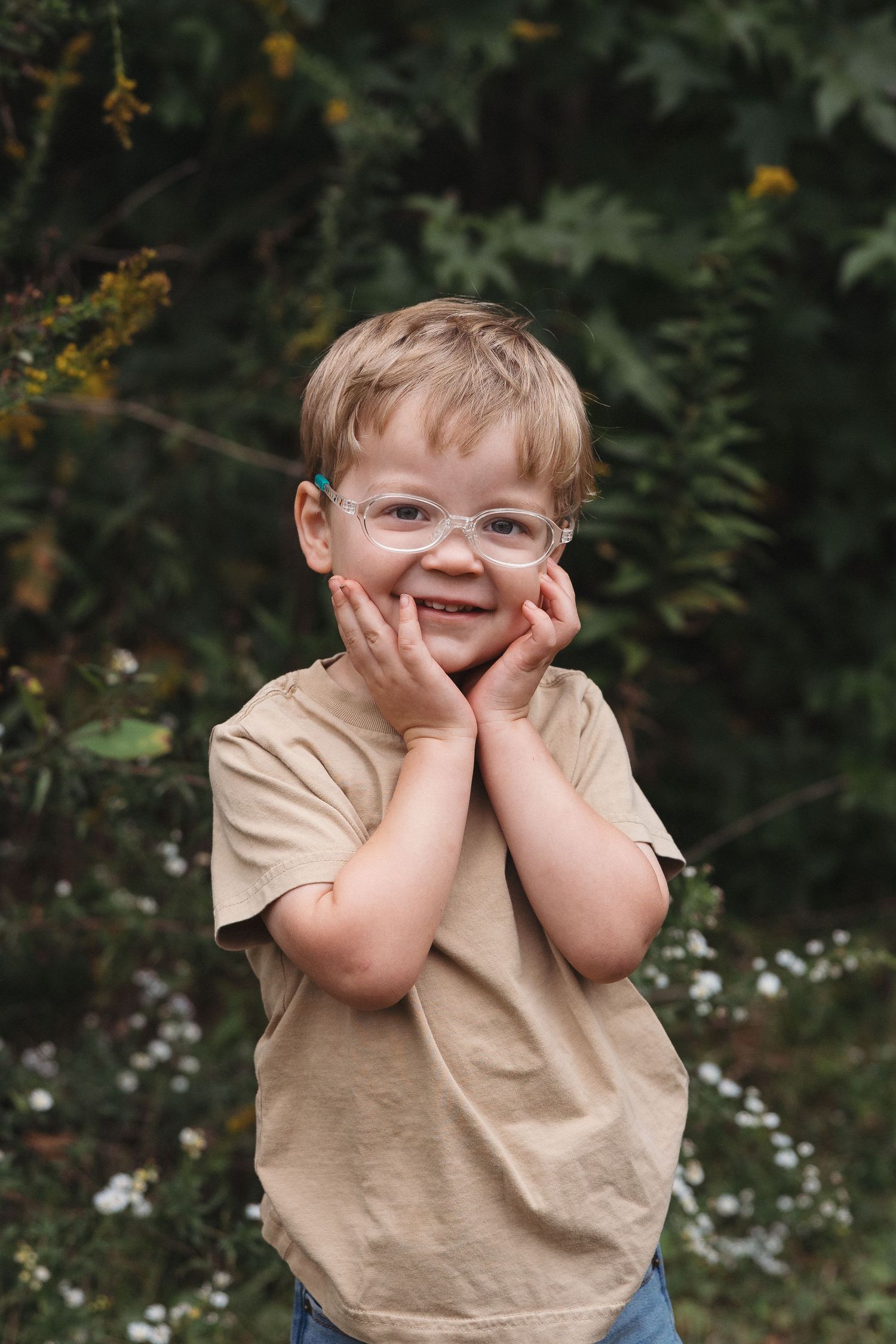 A smiling young boy with glasses and a beige t-shirt poses outdoors with hands on cheeks, surrounded by green foliage.