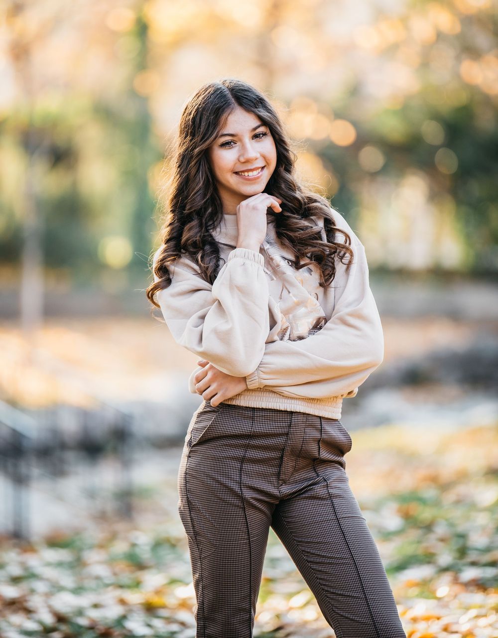 Young woman with dark curly hair smiling outdoors in autumn park, wearing beige top and brown trousers.