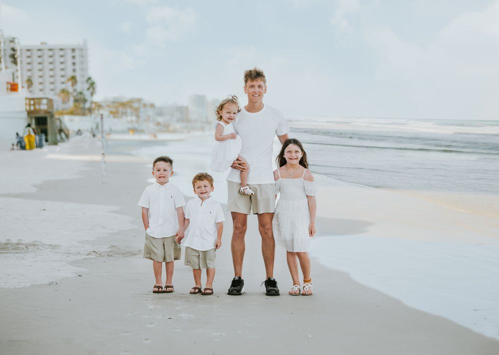Family group in coordinating white and khaki outfits poses together on beach at sunset.