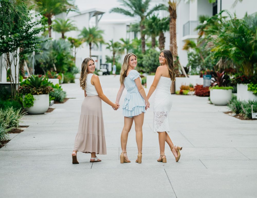 Three friends in summer dresses stroll along a palm-lined walkway at a tropical resort.