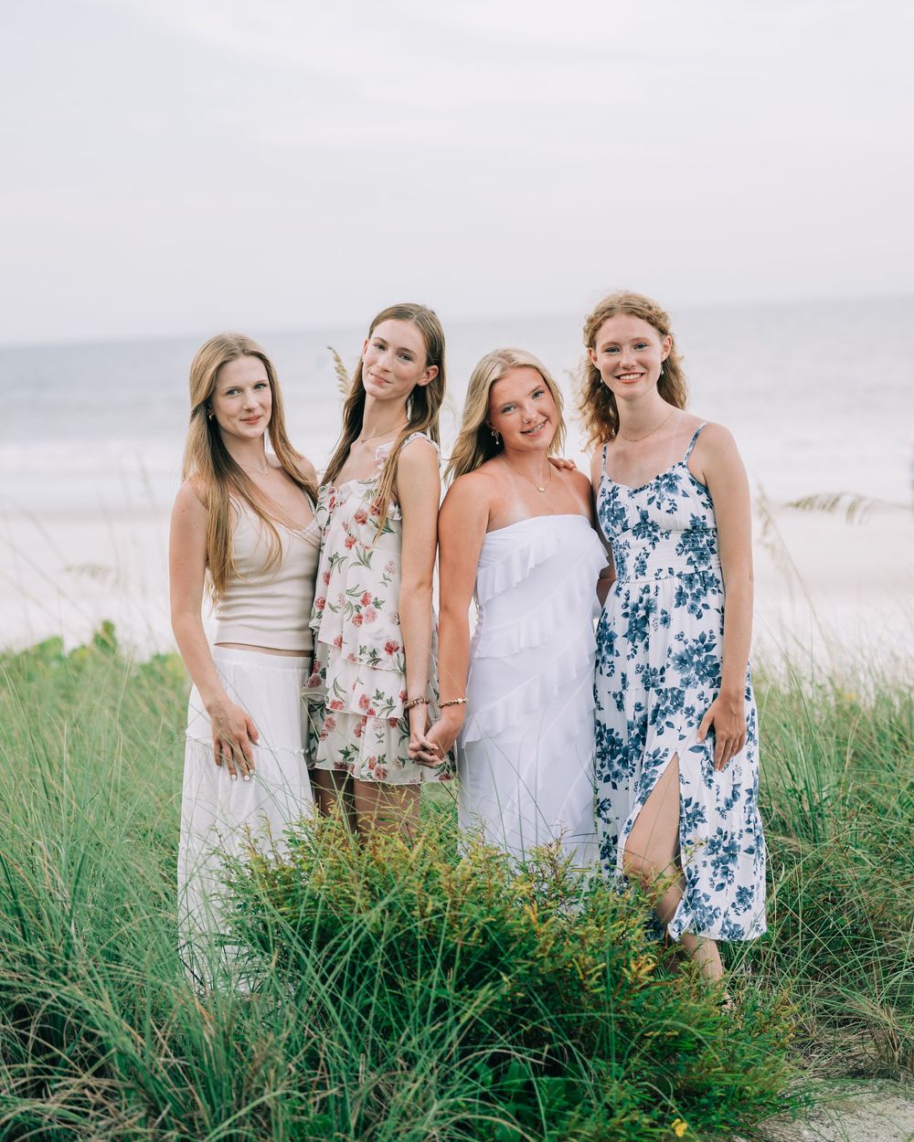 A group of women in summery dresses pose together among beach grass with a coastal background.