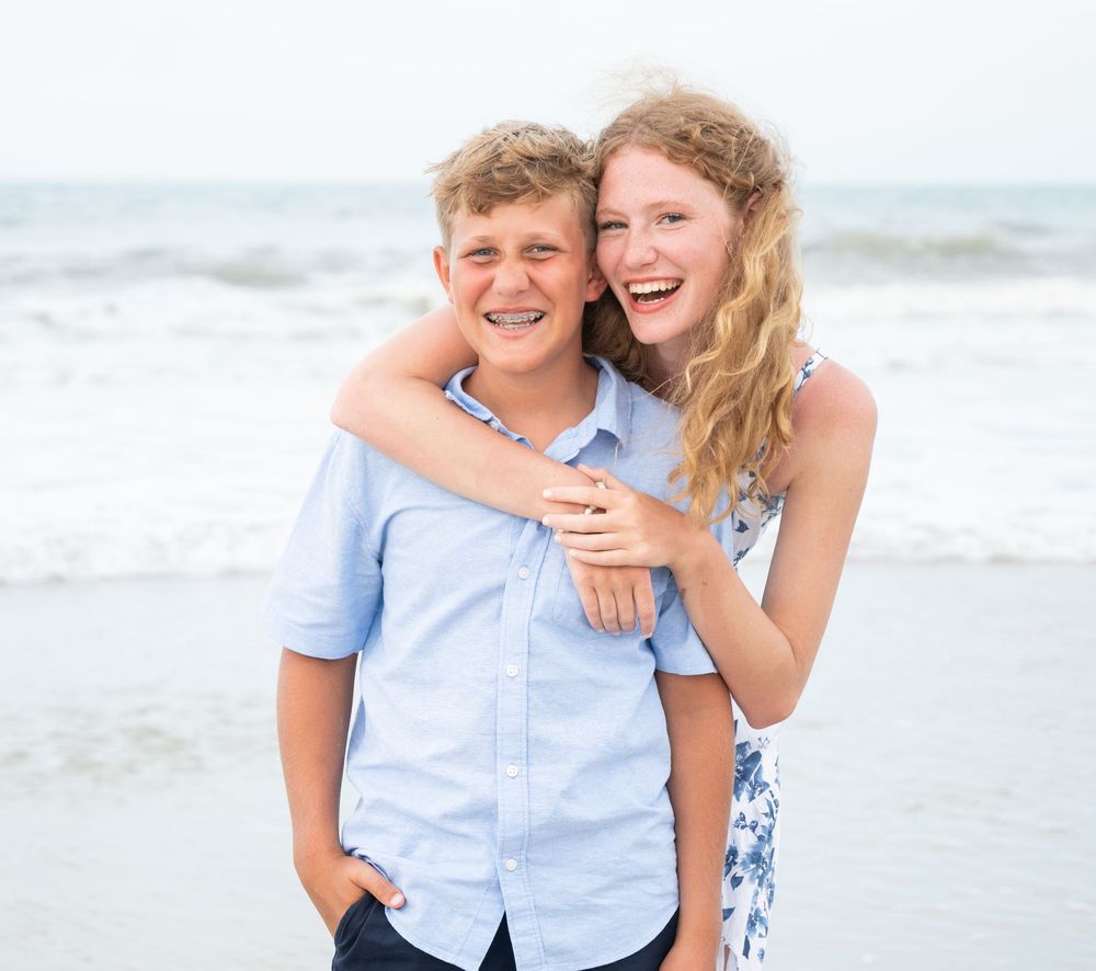 Two people embrace and laugh together while standing on a sunny beach with ocean waves in the background.