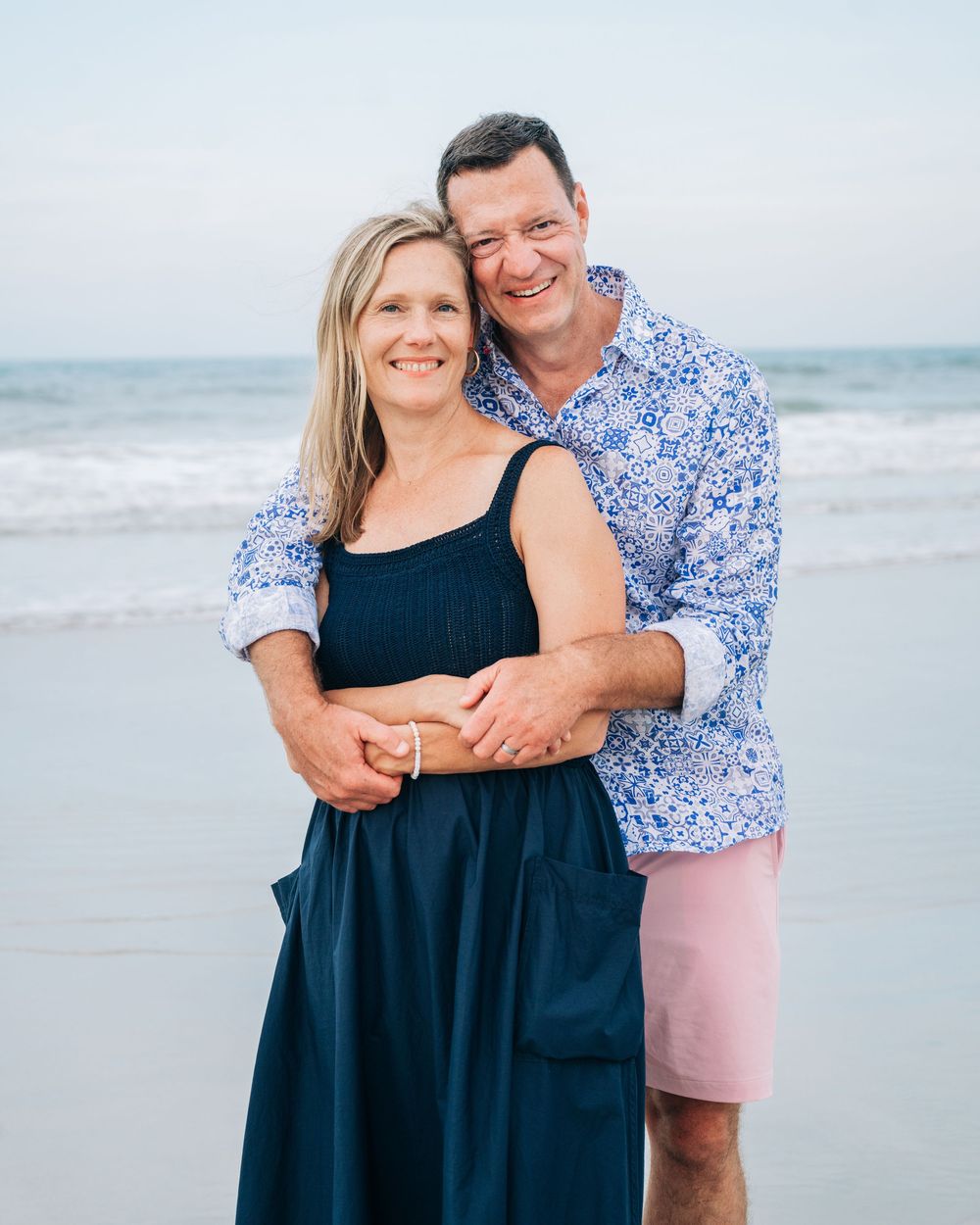 A couple embraces while standing on a beach at sunset, wearing a navy dress and blue patterned shirt with pink shorts.