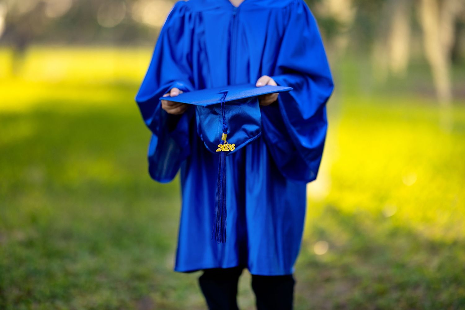 Person in blue graduation gown and cap standing outdoors in a green park setting.