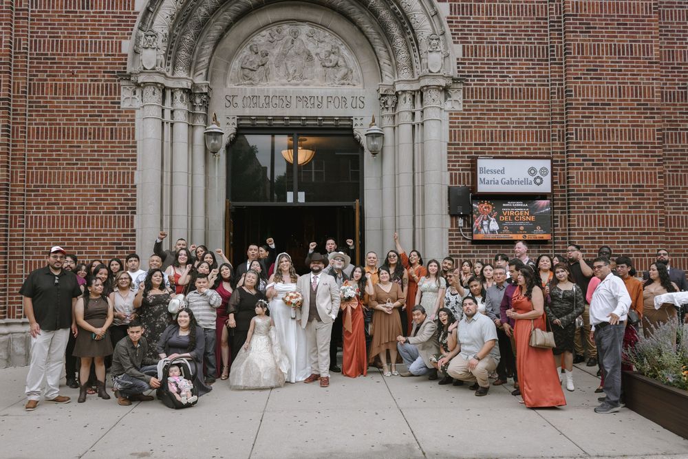 Wedding party poses outside a brick church entrance after a ceremony, with bride and groom surrounded by guests.