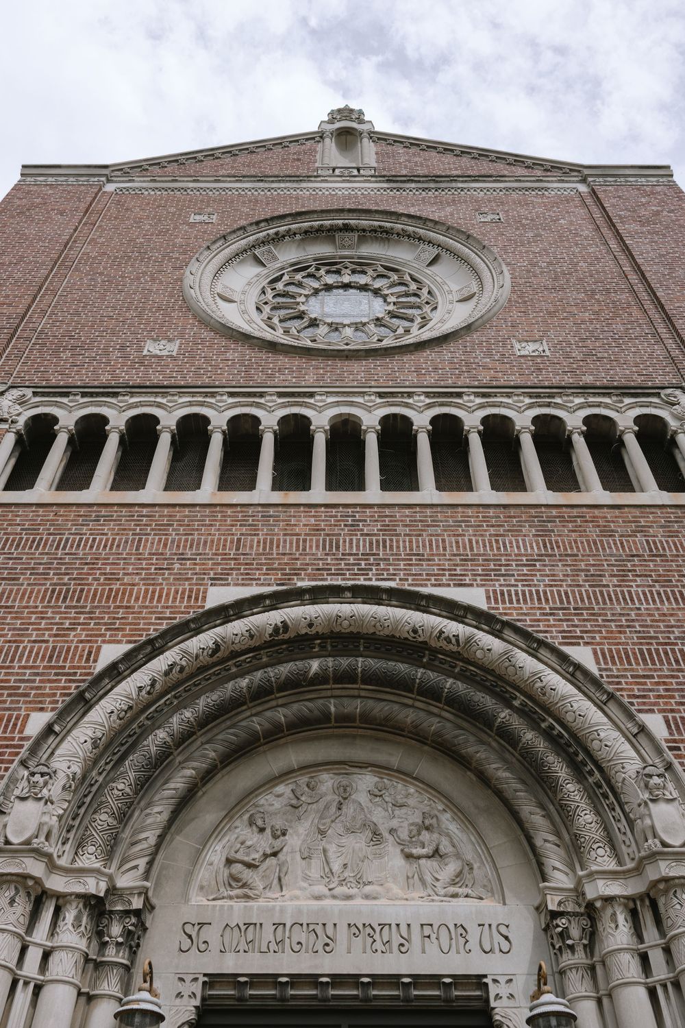 Exterior facade of St. Malachy Catholic Church featuring red brick, rose window, arched entry, and stone inscription.