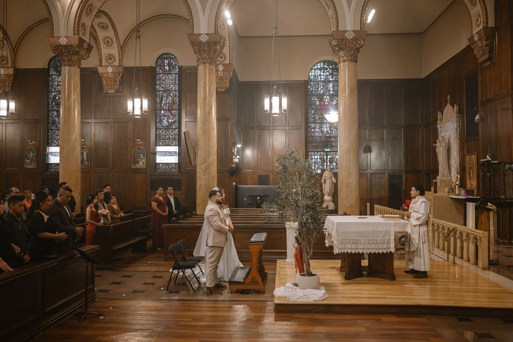 A couple exchanges vows at a Catholic church wedding ceremony with wooden pews, stained glass windows, and an altar.