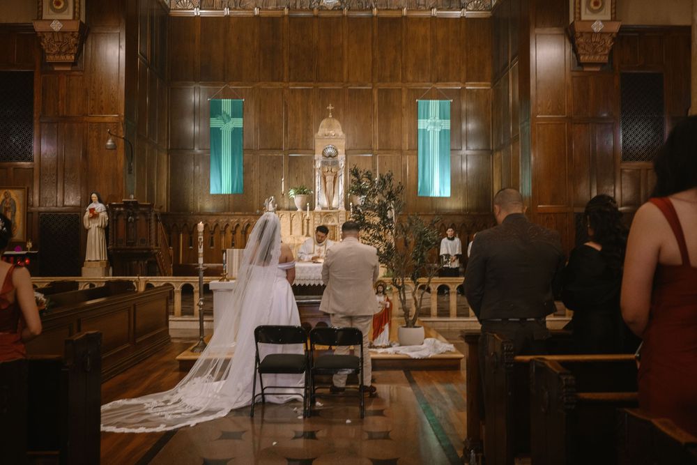 Bride and groom kneeling at altar during Catholic church wedding ceremony with glowing cross windows.