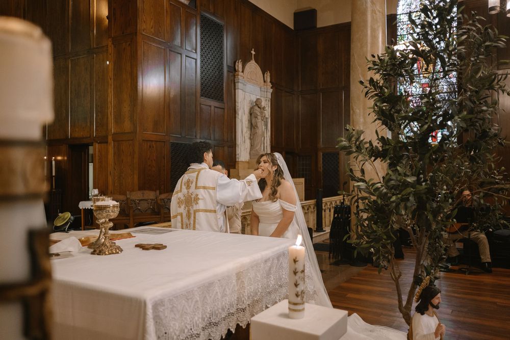 Bride and groom at church altar during wedding ceremony with candles and olive tree in elegant wood-paneled chapel.