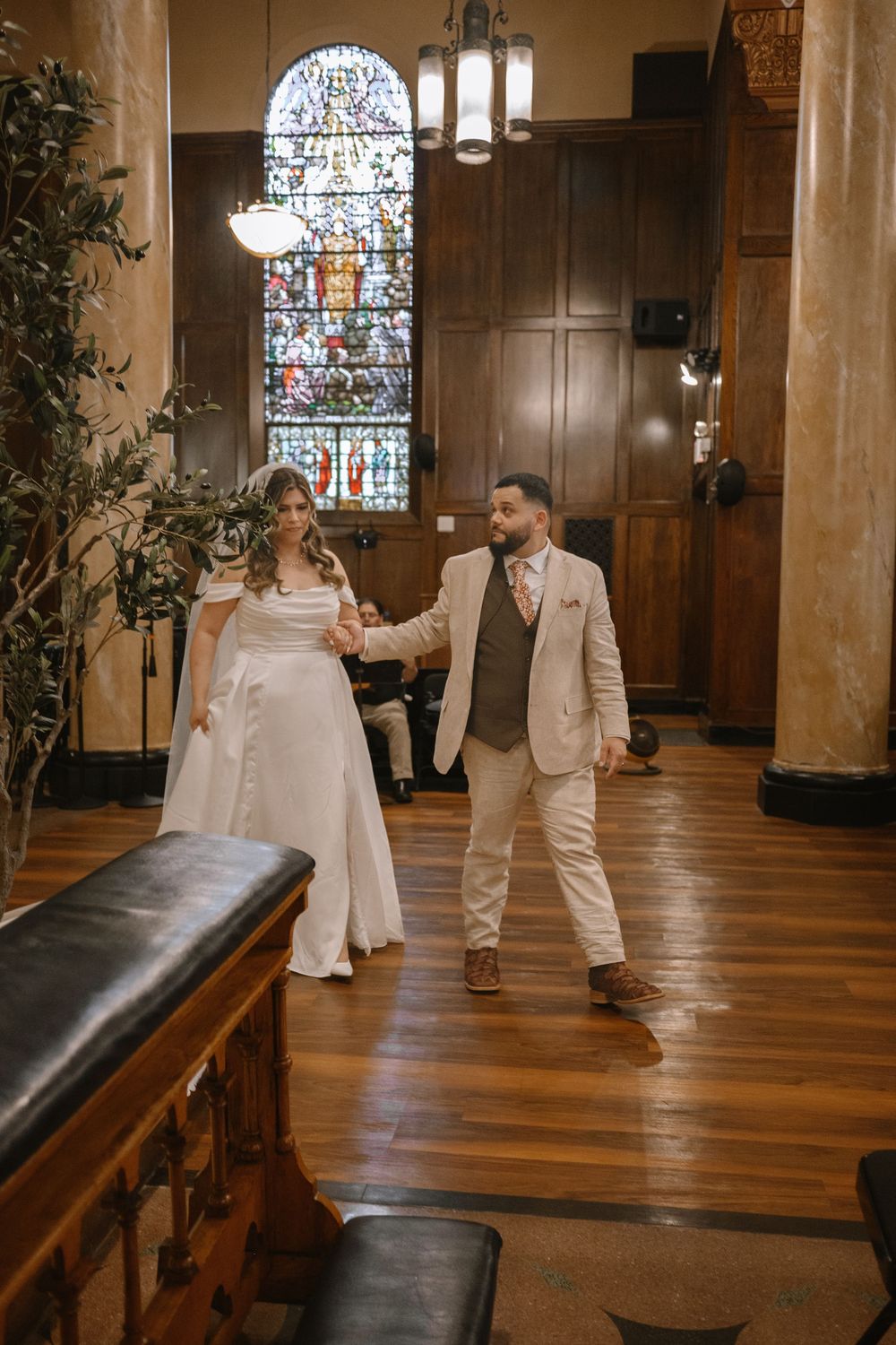 Bride and groom walk hand in hand through an elegant wood-paneled hall with stained glass windows on their wedding day.