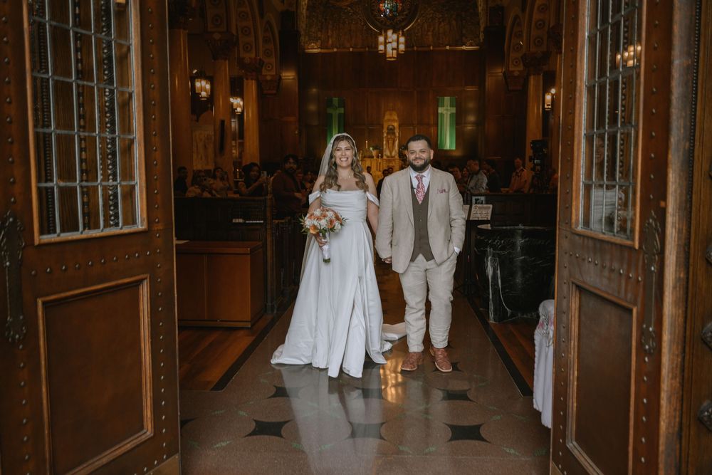Bride in white gown and groom in tan suit walk down church aisle after wedding ceremony.