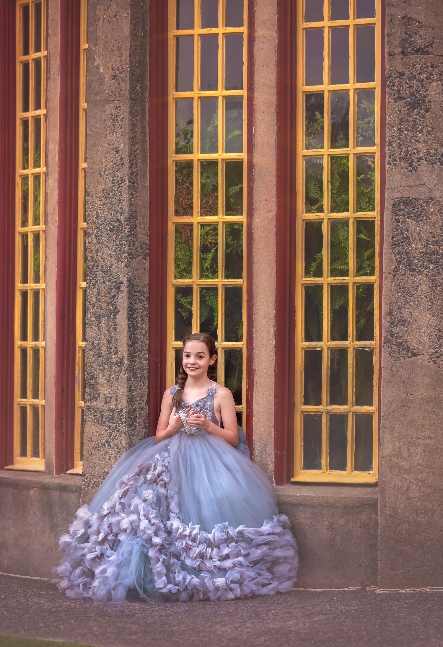 A young girl in a stunning blue ball gown poses gracefully in front of yellow-framed windows at an elegant building.