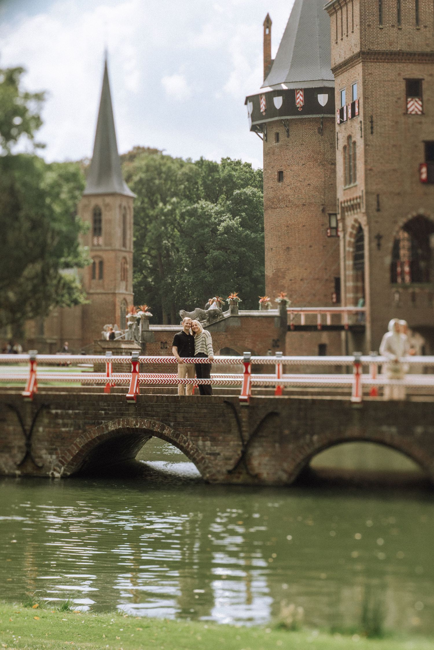 Couple embracing on a stone arch bridge over calm water beside a historic brick castle De Haar with towers and a church spire.