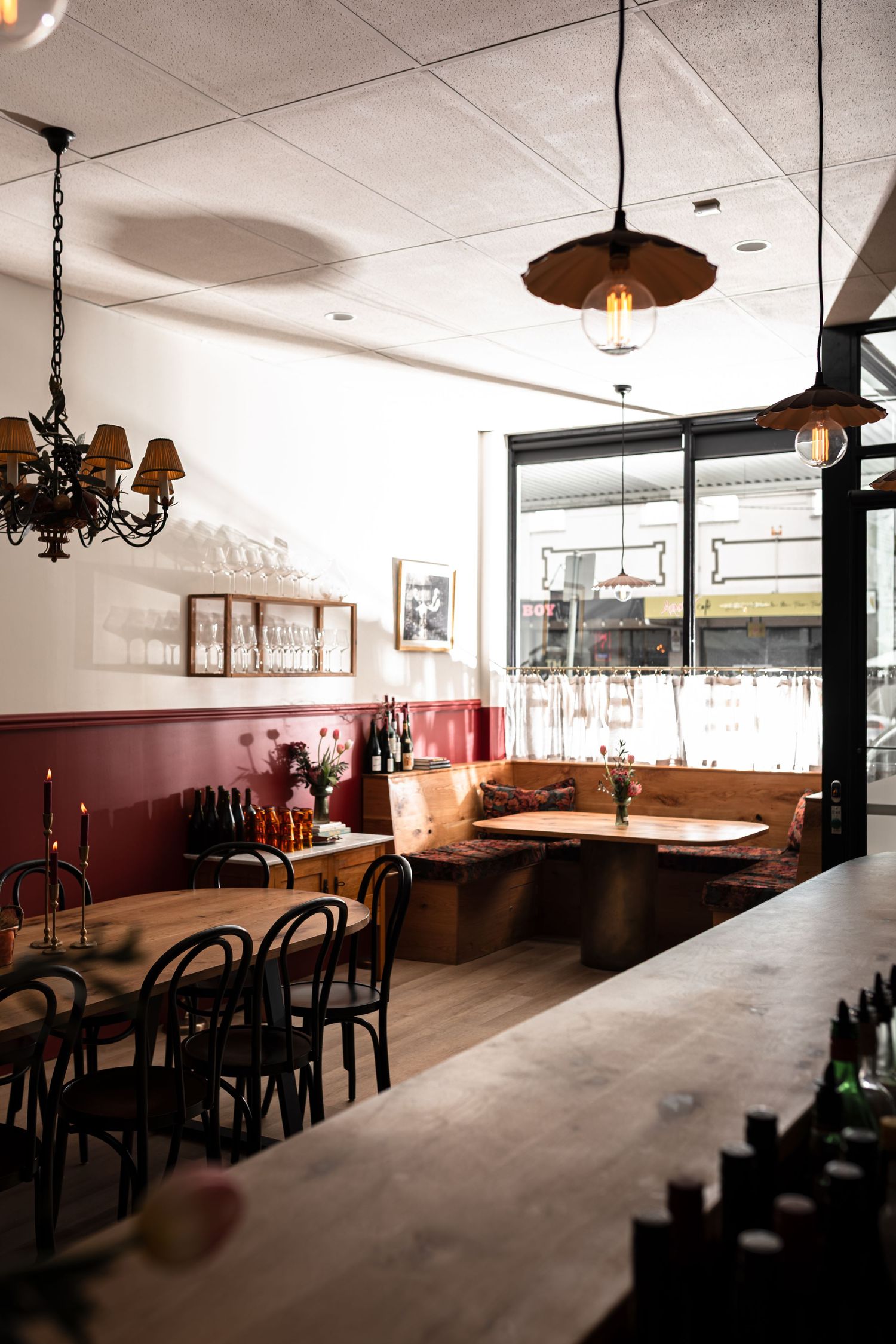 Cozy restaurant interior with burgundy wainscoting, wooden bentwood chairs, a corner banquette, pendant lamps and a wine bottle bar.