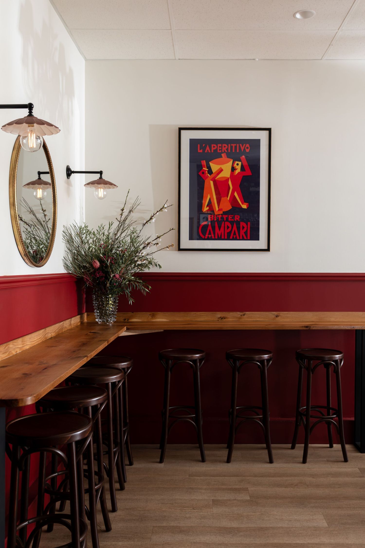 Cozy bar corner with a wooden counter, black stools, red wainscoting, a vintage Campari poster, round mirror and eucalyptus in a vase.