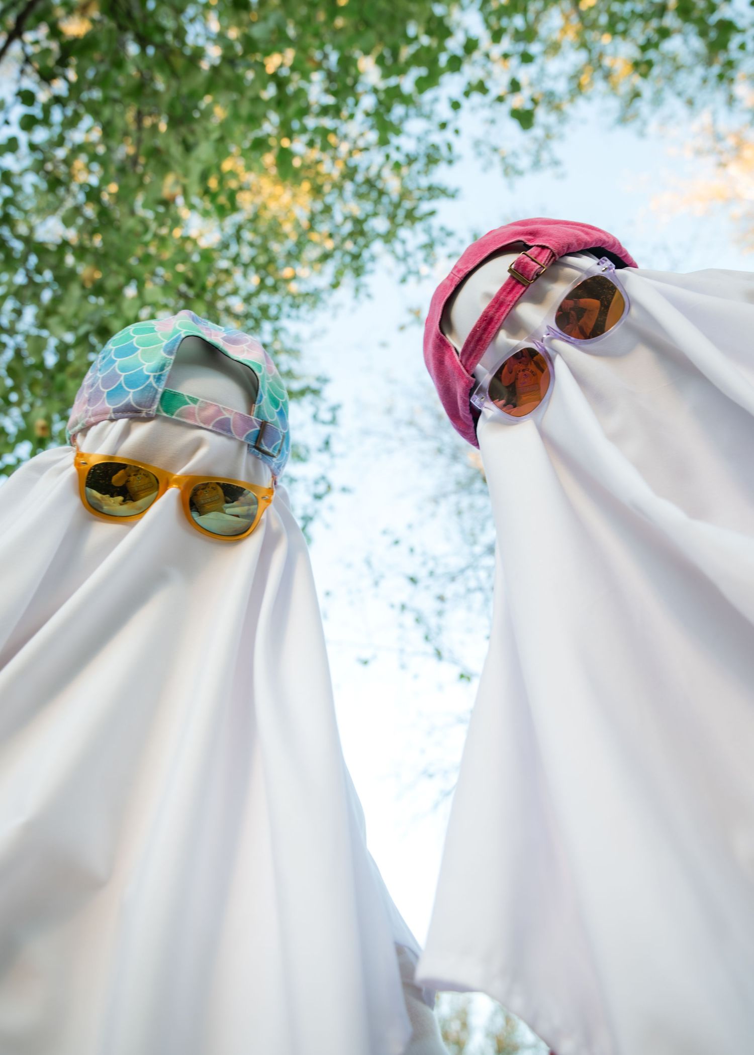 Two people in white ghost sheet costumes with colorful caps and mirrored sunglasses stand under green trees, seen from a low angle.