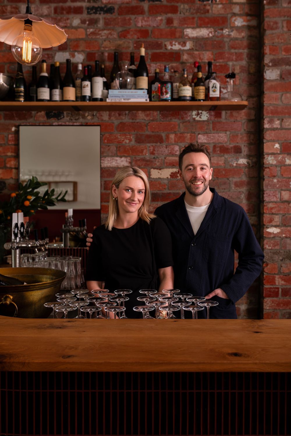 Smiling man and woman stand behind a wooden bar with rows of wine glasses, a bottle shelf and an exposed brick wall behind them.