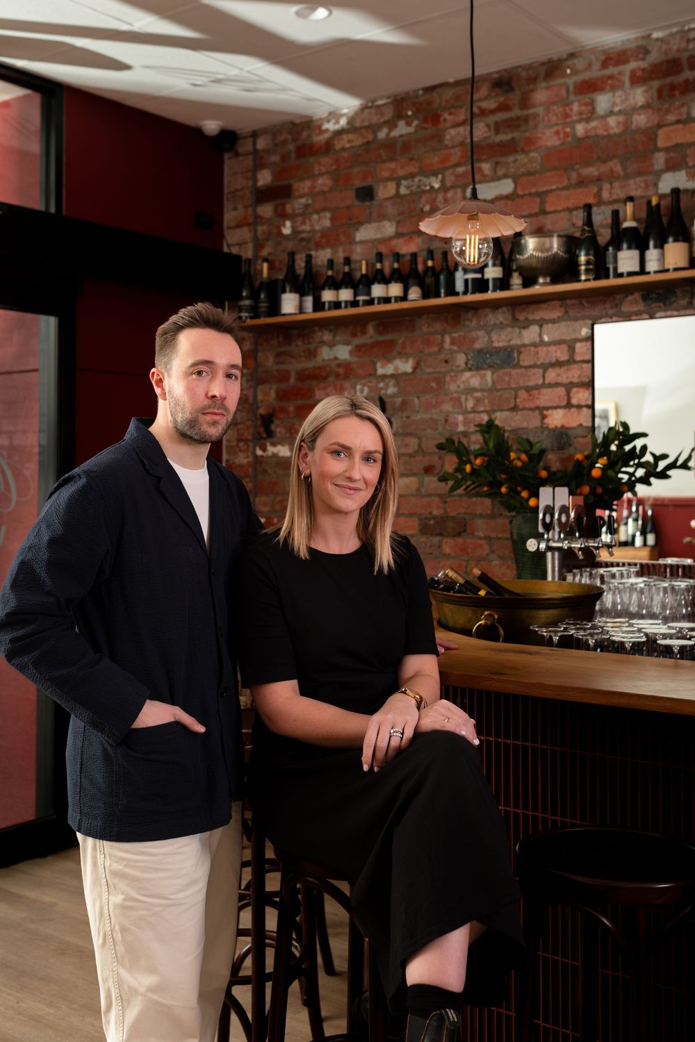 A man and woman pose together at a wine bar with exposed brick walls, a wooden counter, and shelves of wine bottles behind them.