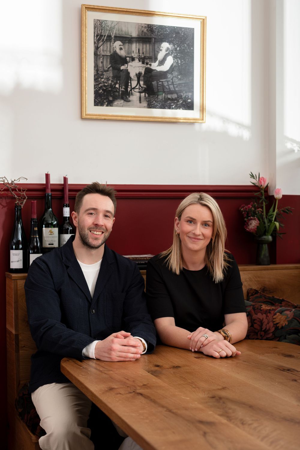 A smiling man and a blonde woman sit side by side at a wooden table in a cozy restaurant with wine bottles, tulips and a framed photo.