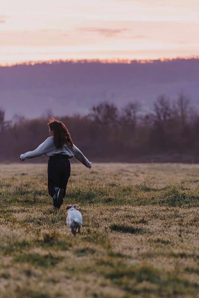 Woman in a cozy sweater runs with arms outstretched across a grassy field toward a small dog at a pink sunset.