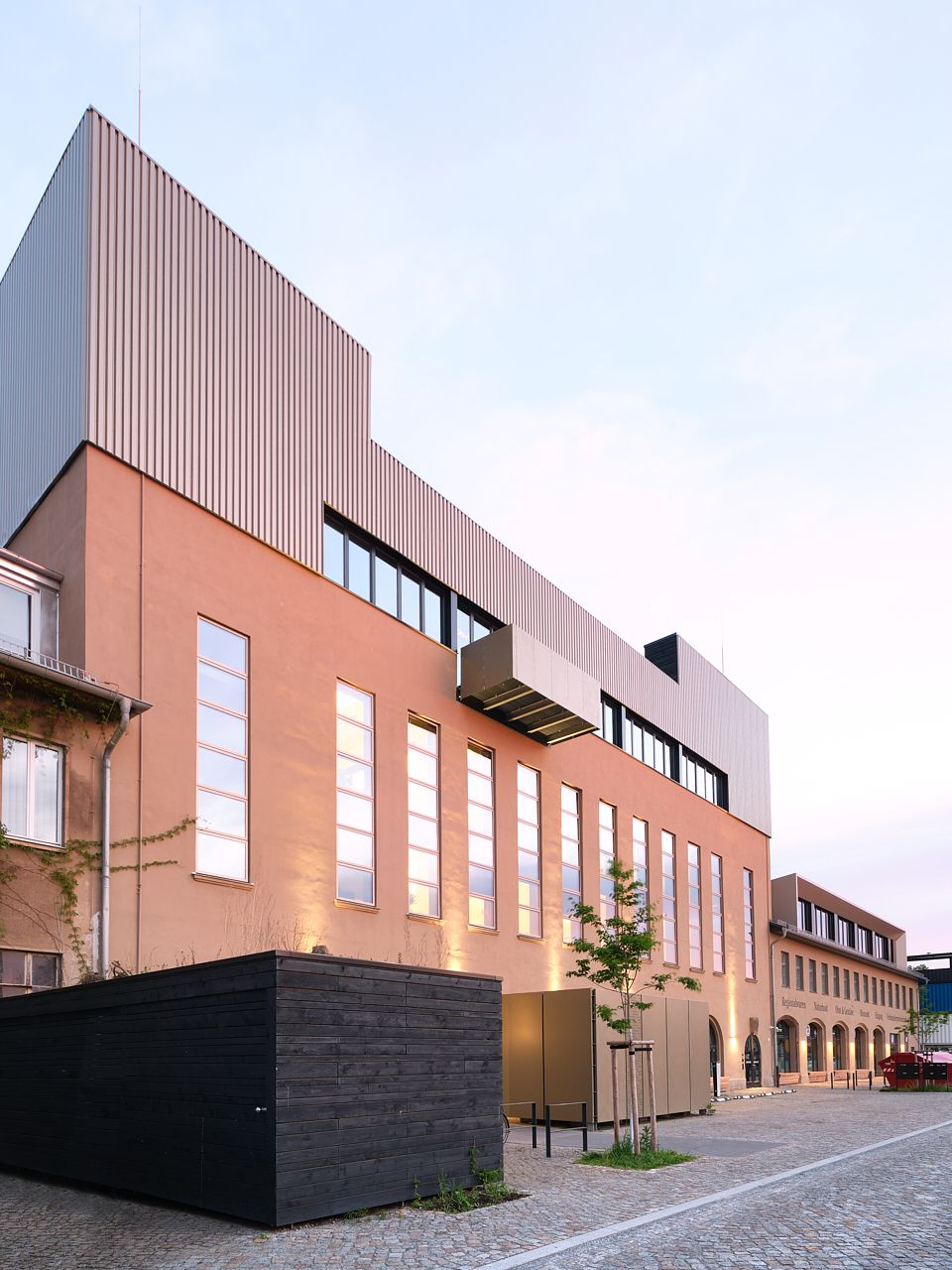 Modern brick building with tall windows and a corrugated metal upper floor beside a cobblestone plaza at dusk.