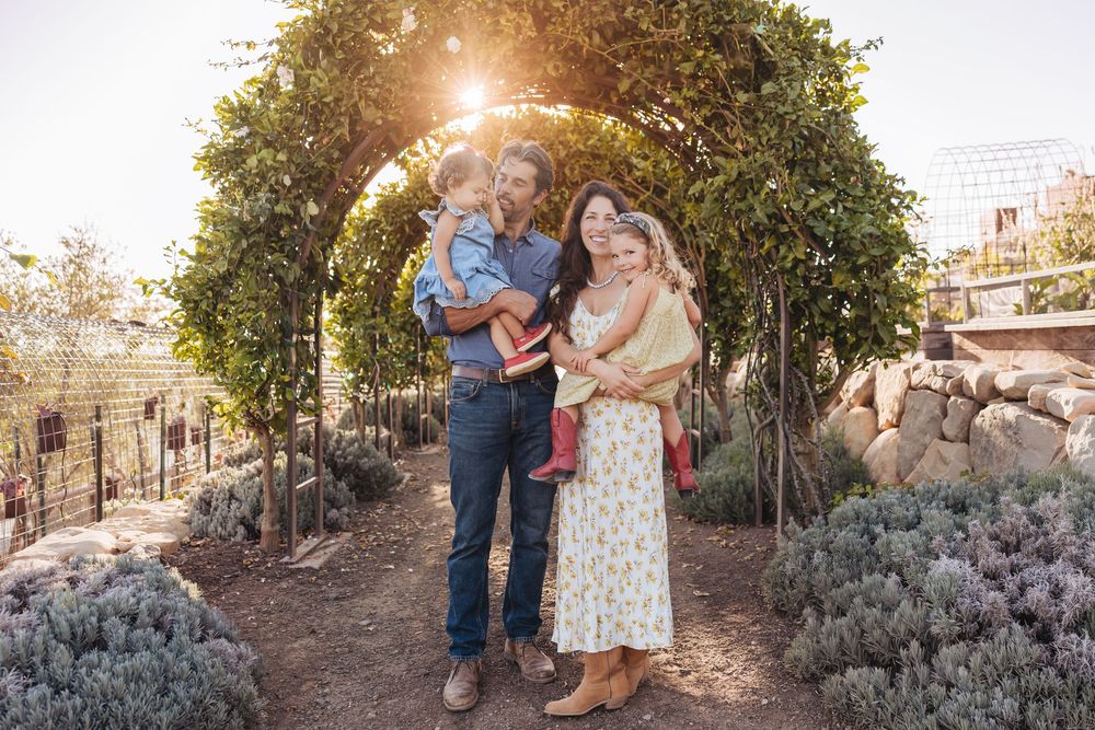 A smiling family of four poses beneath a leafy garden arch with sunlight streaming through the greenery.