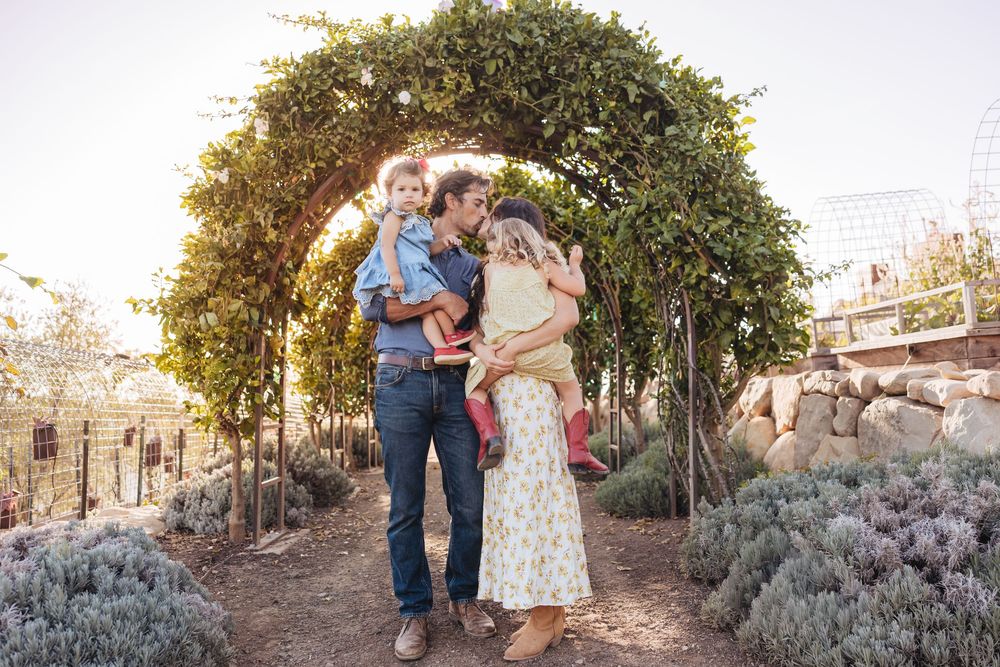 A father and mother hug and hold two young daughters beneath a leafy garden archway surrounded by lavender and stone walls.