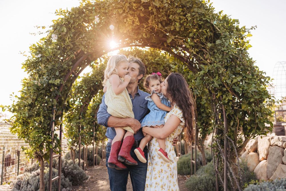 A smiling couple holds two young daughters beneath a leafy garden arch as warm golden sunlight shines through.