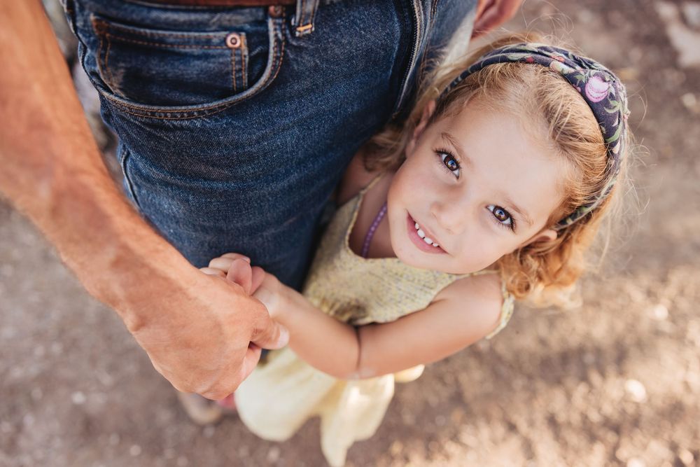 A smiling little girl in a floral dress and headband holds a man's hand as he stands beside her in jeans.