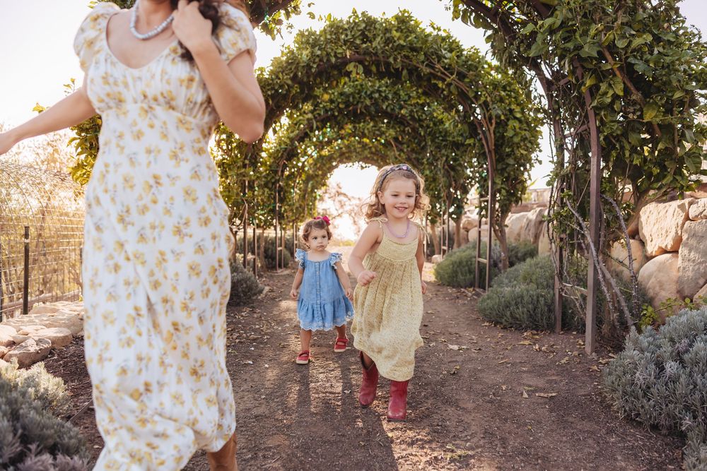 Two young girls in dresses and red boots run along a garden path under a leafy arch as their mother walks nearby.