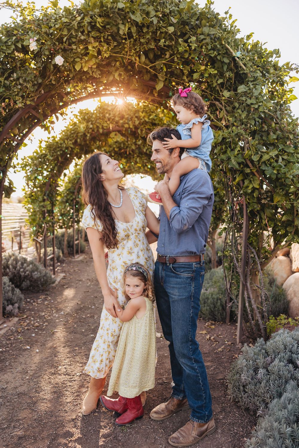 A smiling family of four stands beneath a leafy garden archway at sunset, with a toddler riding on her father's shoulders.