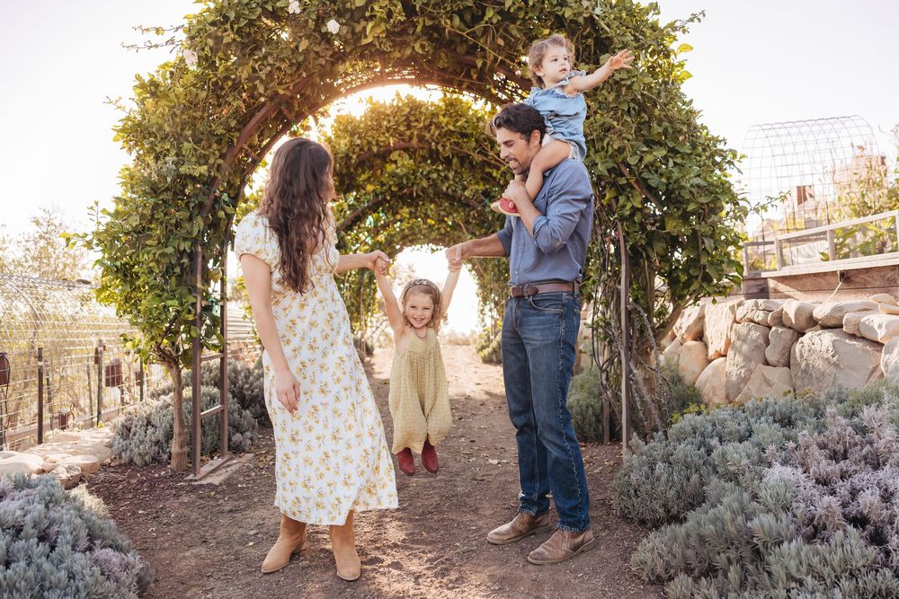 Parents lift a laughing girl by the hands under a vine-covered arch while a toddler perches on dad's shoulders.