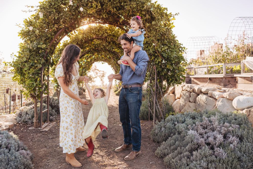 A joyful family plays beneath a garden arch as parents swing a toddler girl while another child rides on dad's shoulders.