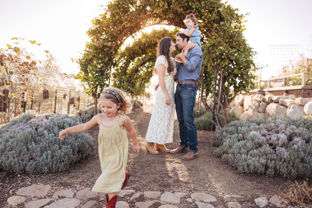 A young girl runs down a stone garden path toward the camera while her parents and toddler stand beneath a greenery arch.
