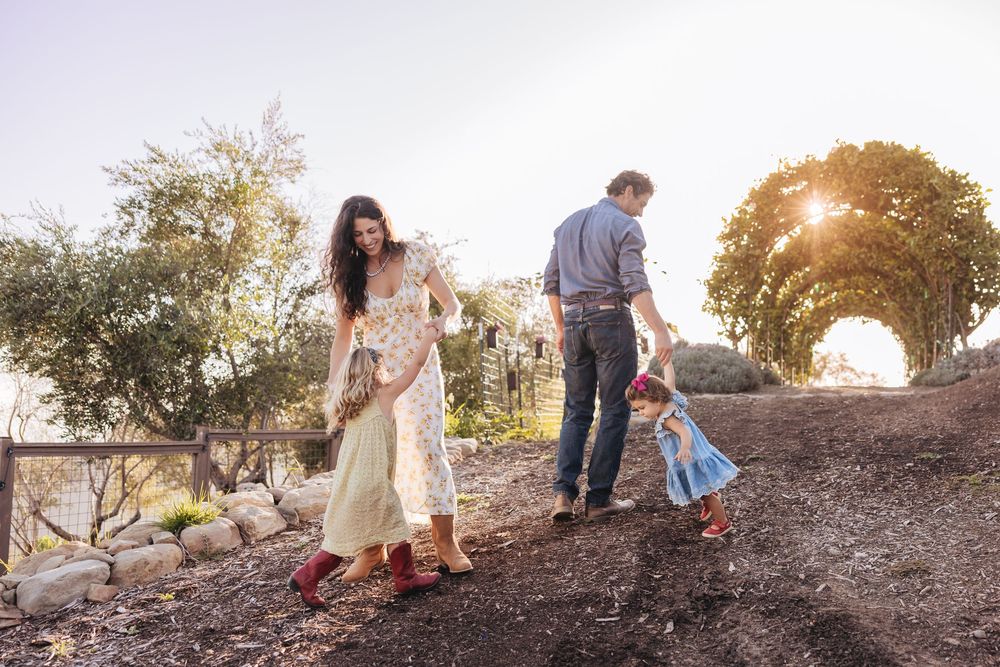 A family walks along a garden path at sunset as a toddler in a blue dress follows her parents toward a glowing arch.