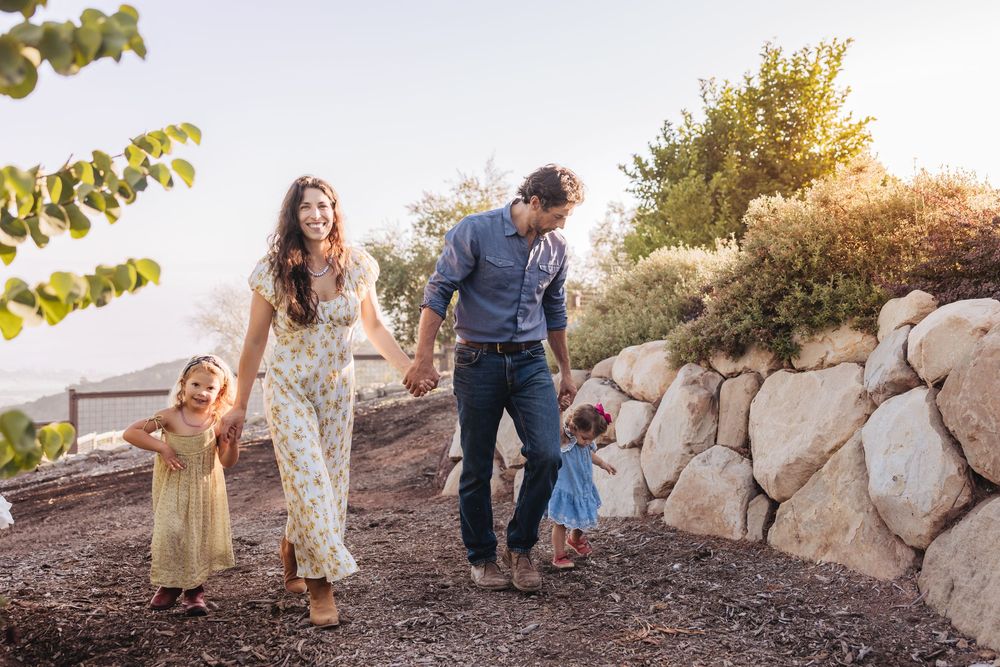 A happy family of four holds hands and strolls along a rocky garden path in warm golden light.