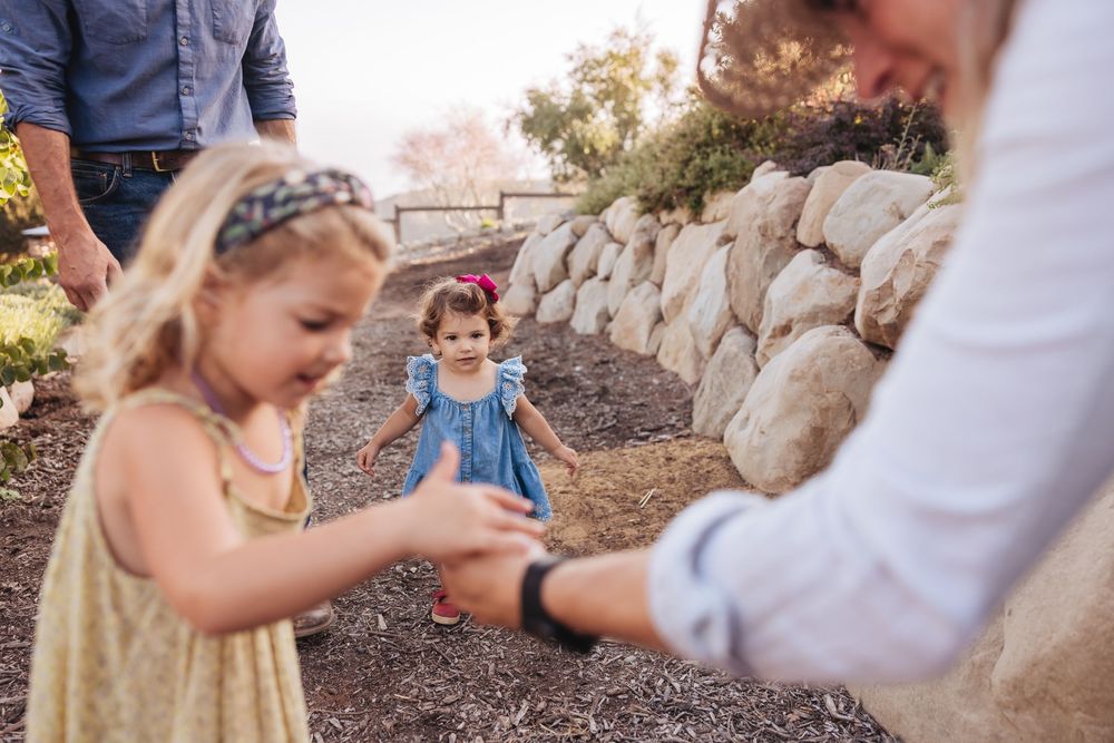 A toddler in a blue dress and a girl in a yellow dress reach for a parent's hand beside large garden boulders.