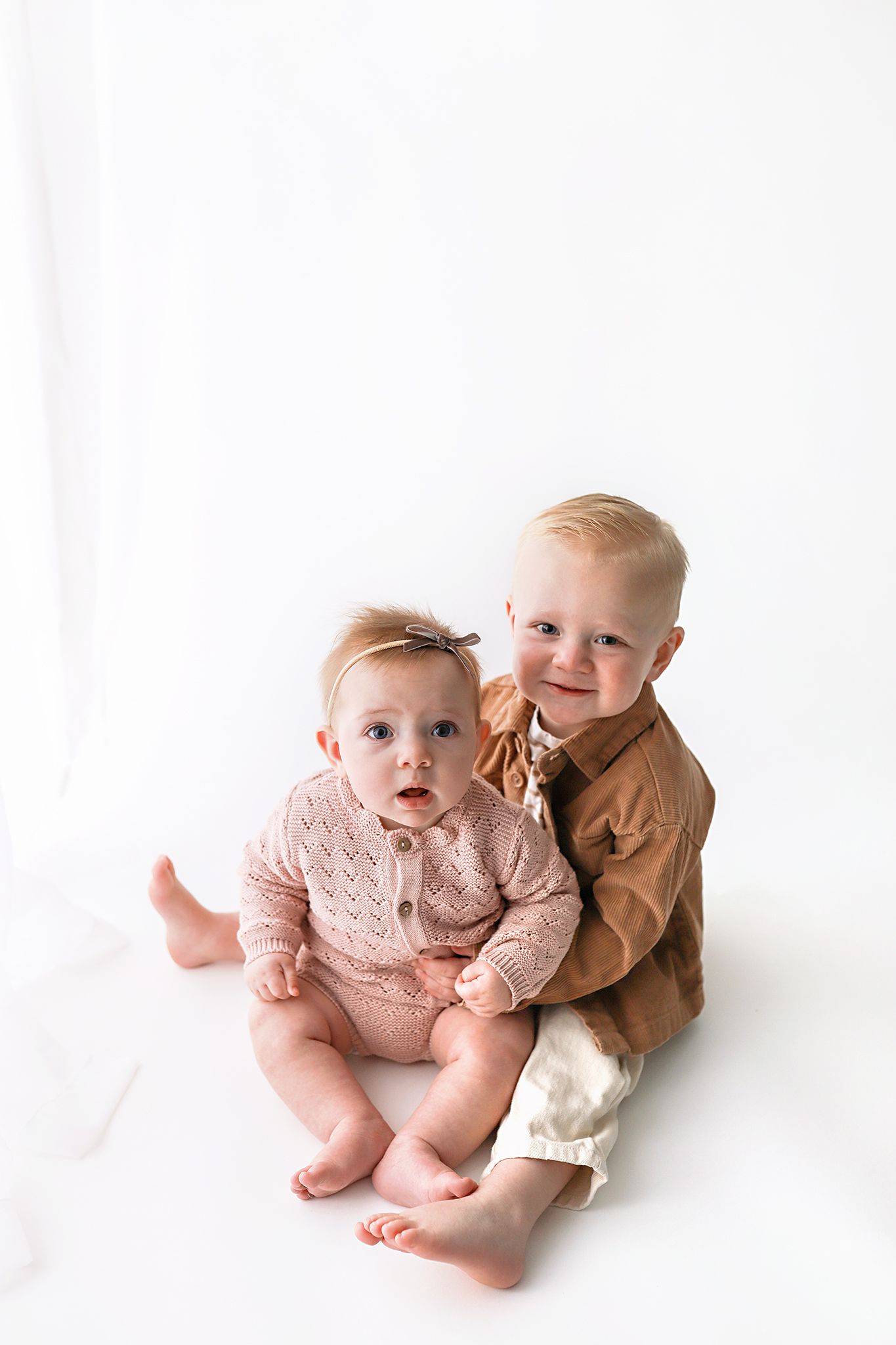 Two smiling toddlers sit close together on a white studio background, one in a pink knit outfit with a bow and one in a tan jacket.