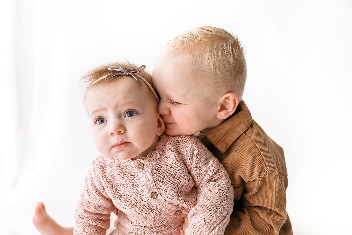 A toddler nuzzles and kisses the cheek of a baby girl against a white background. Utah baby photographer. Utah County photographer