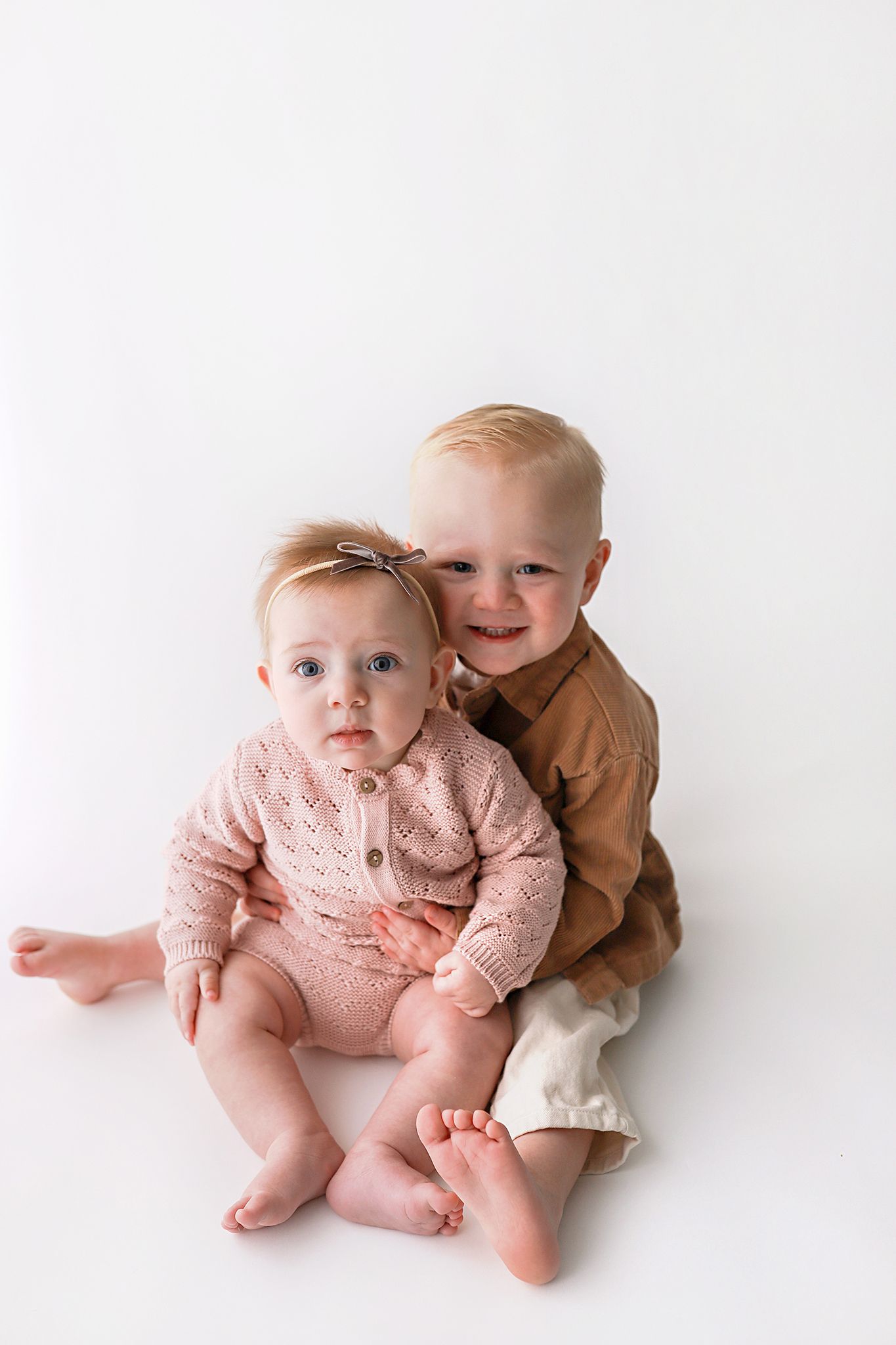 Smiling toddler boy in a tan shirt hugs a baby girl in a pink knit romper and bow headband on a white studio background.
