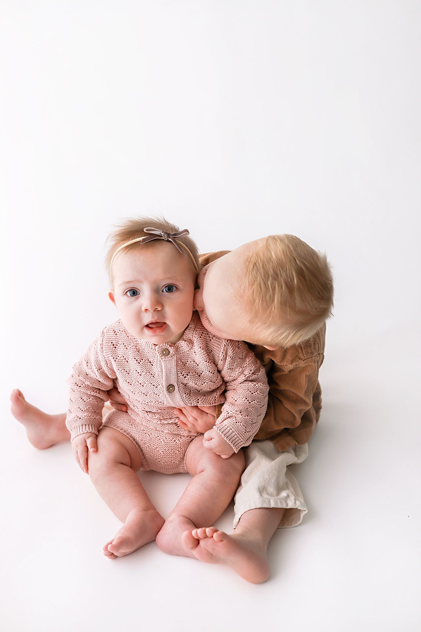 Baby girl in a pink knit romper and bow headband sits on a white background while a toddler sibling kisses her cheek.