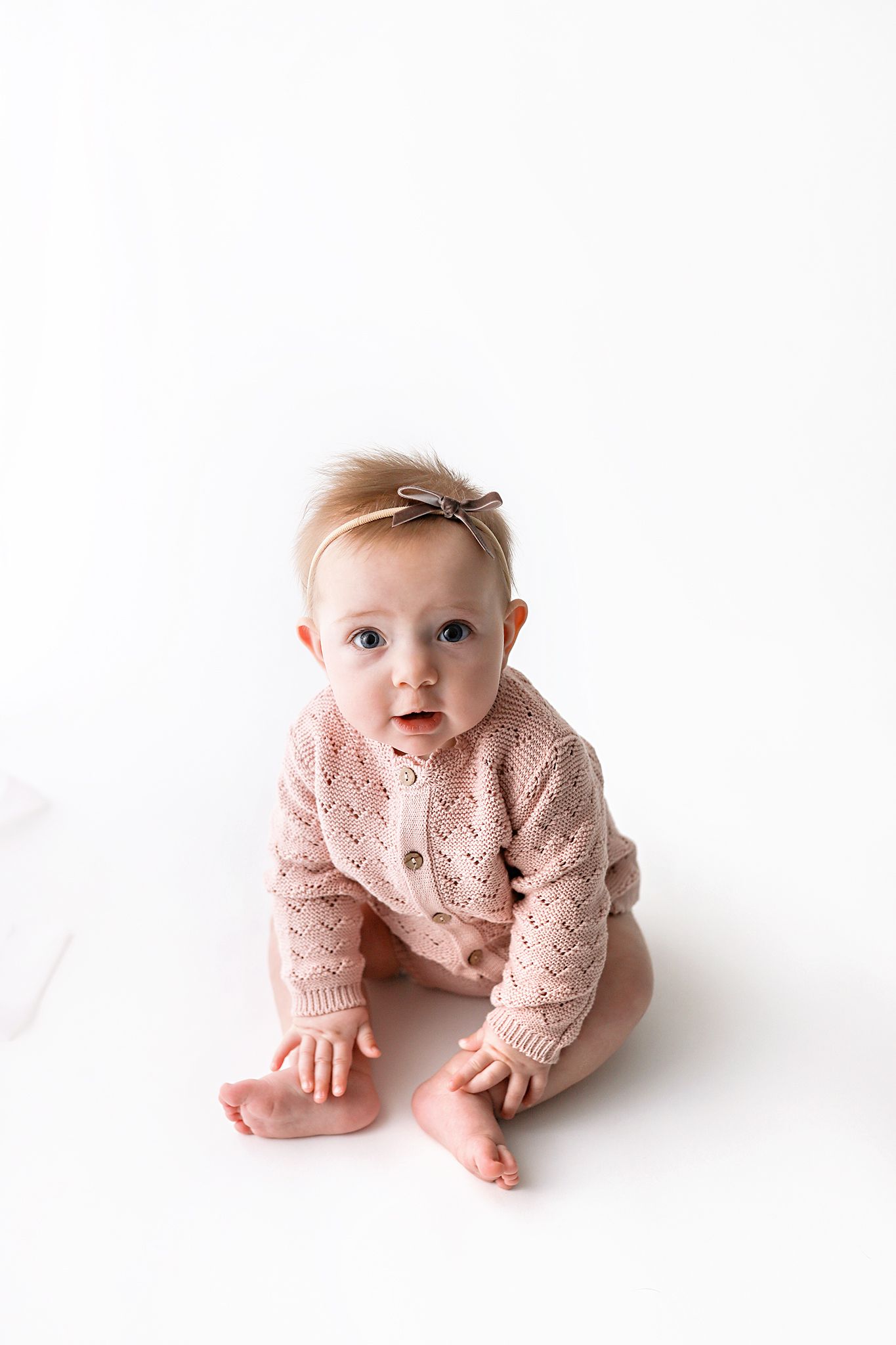 Baby girl in a pink knit cardigan romper and bow headband sits and looks up at the camera against a bright white studio background.
