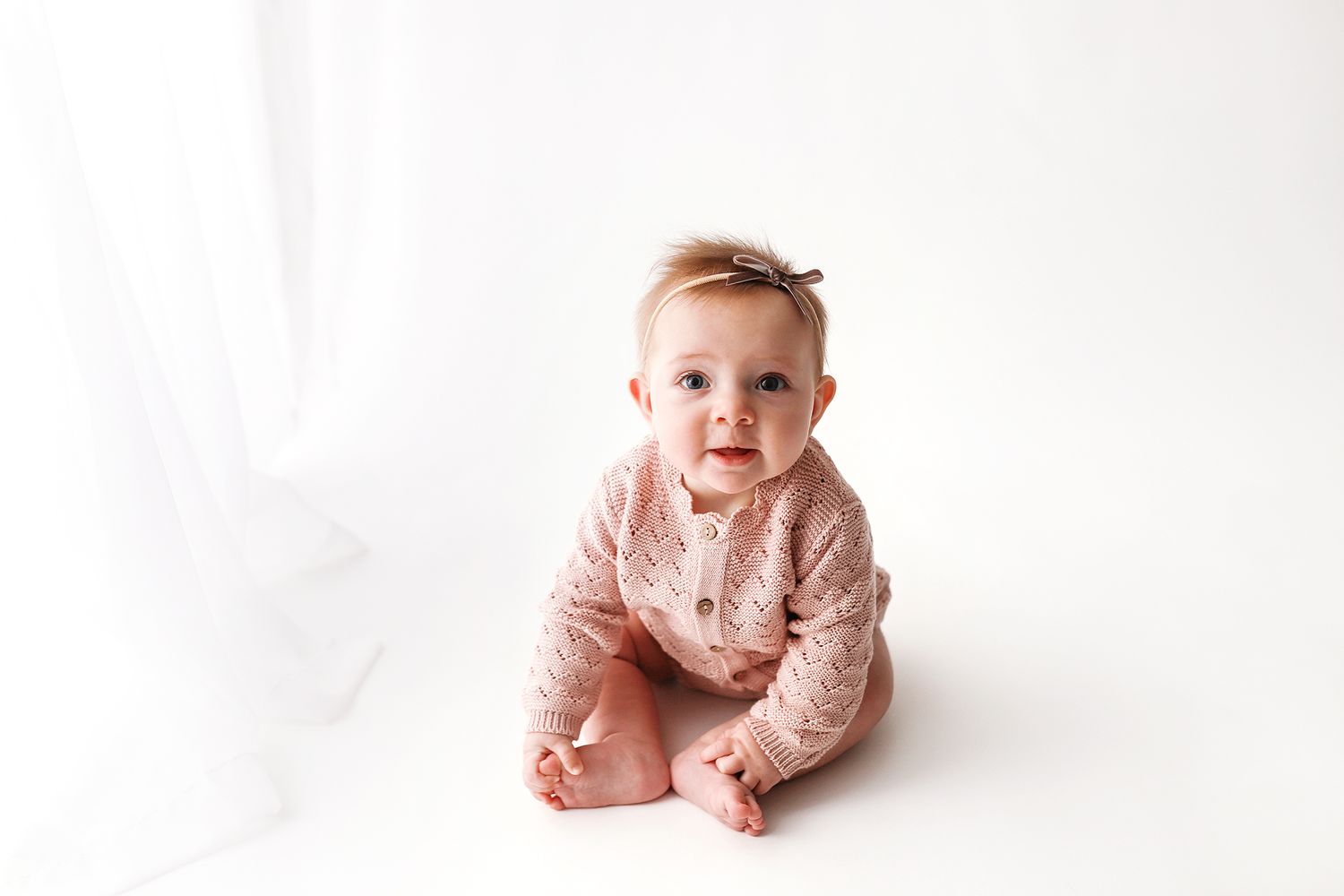 Smiling baby girl in a pink knit outfit and bow headband sitting on a white studio backdrop with soft sheer curtains. Utah baby photographer