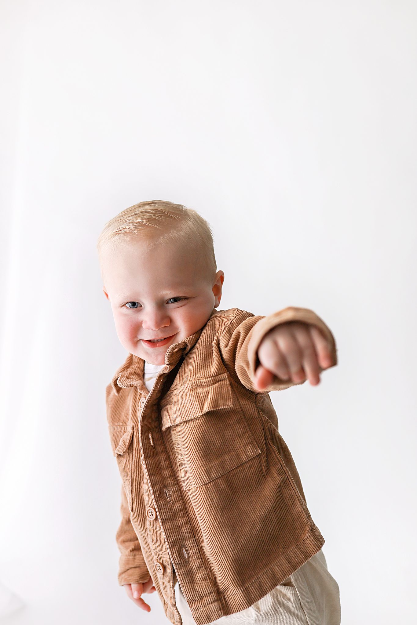 Smiling blond toddler in a tan corduroy button-up jacket reaches one arm toward the camera against a bright white studio background.