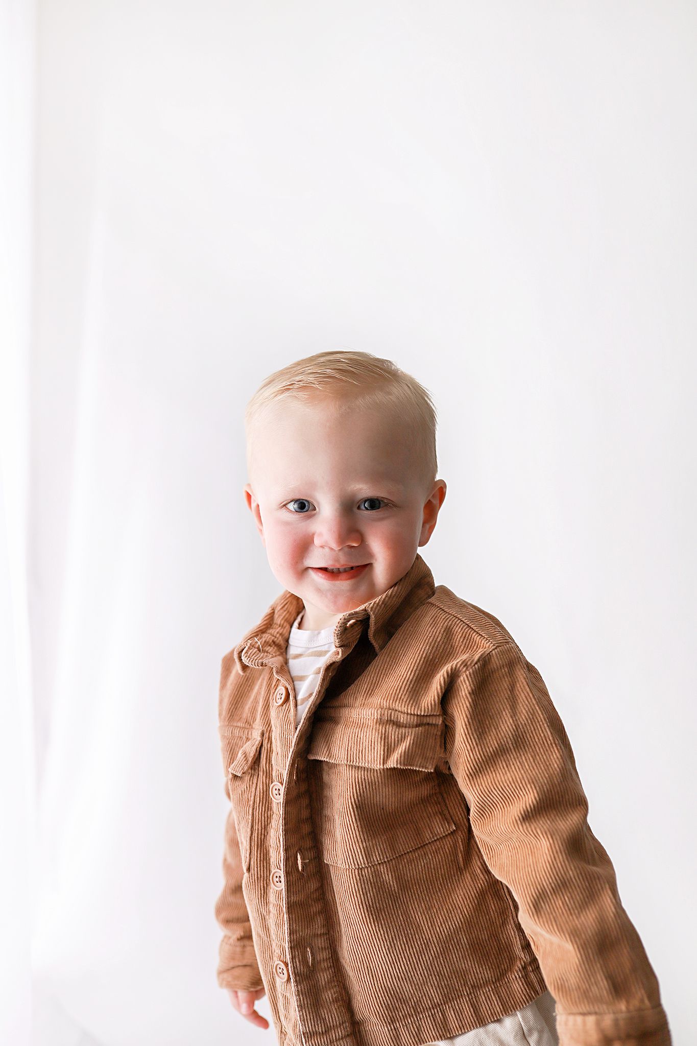 Smiling blond toddler with blue eyes wearing a tan corduroy button-up jacket over a white shirt against a soft white backdrop.